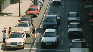 Foto: Richtungsfahrbahn mit 3 gleich breiten Fahrstreifen - Radfahrer zwischen parkenden und fahrenden Autos. 